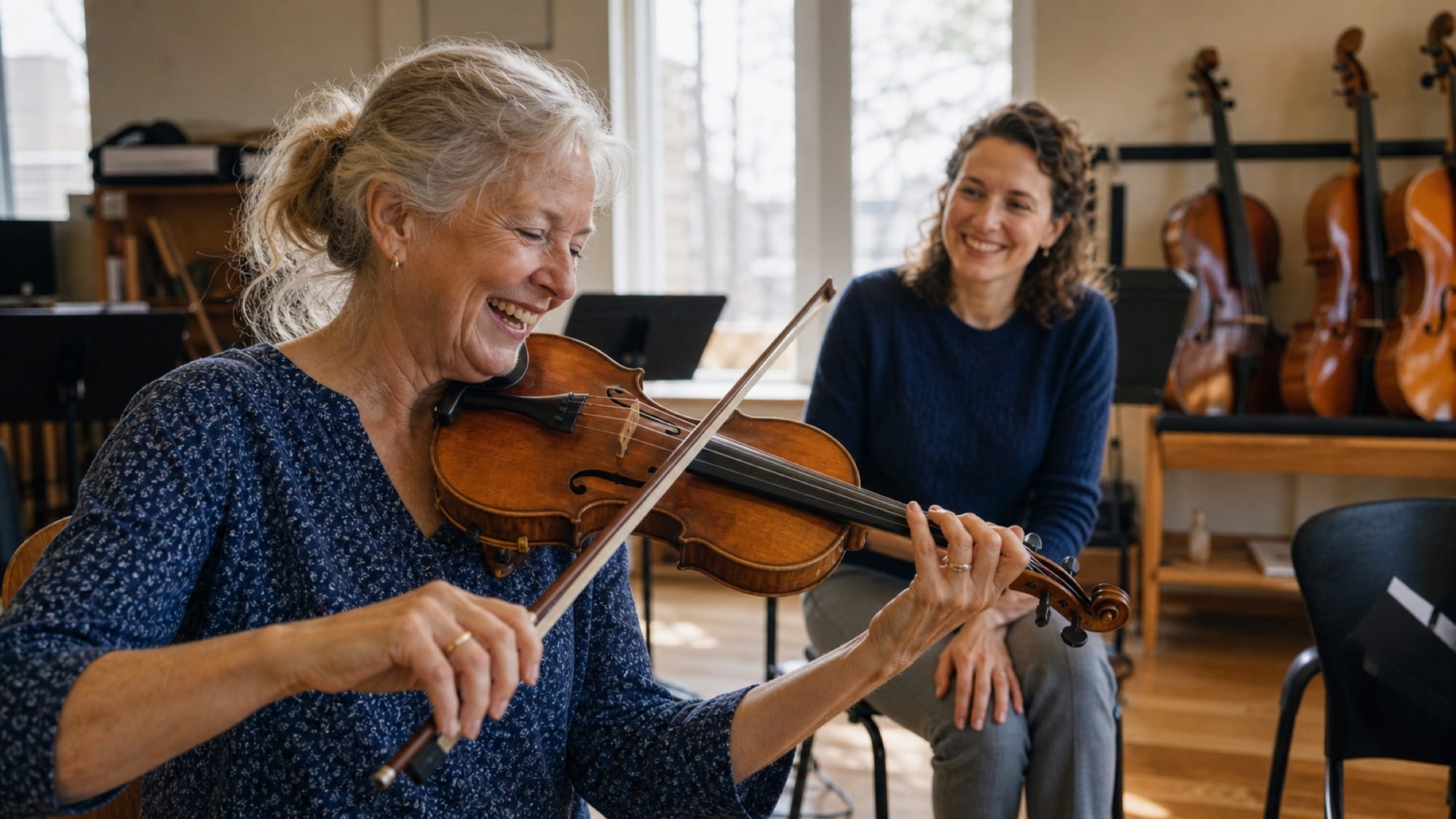 An older woman plays violin in a sunlit room while a smiling woman watches, cellos lined up behind them.